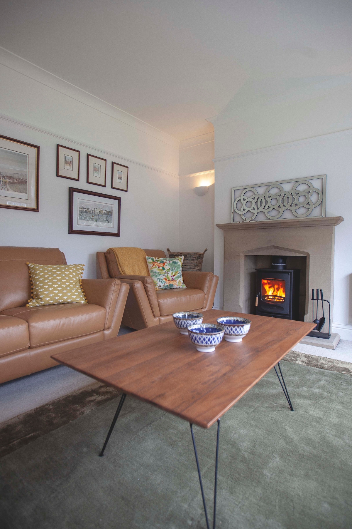 A comfortable lounge area is presented, featuring two tan leather armchairs beside a wooden coffee table. A soft green rug complements the space, while a modern fireplace adds warmth. Stylish wall art and decorative cushions enhance the inviting atmosphere.