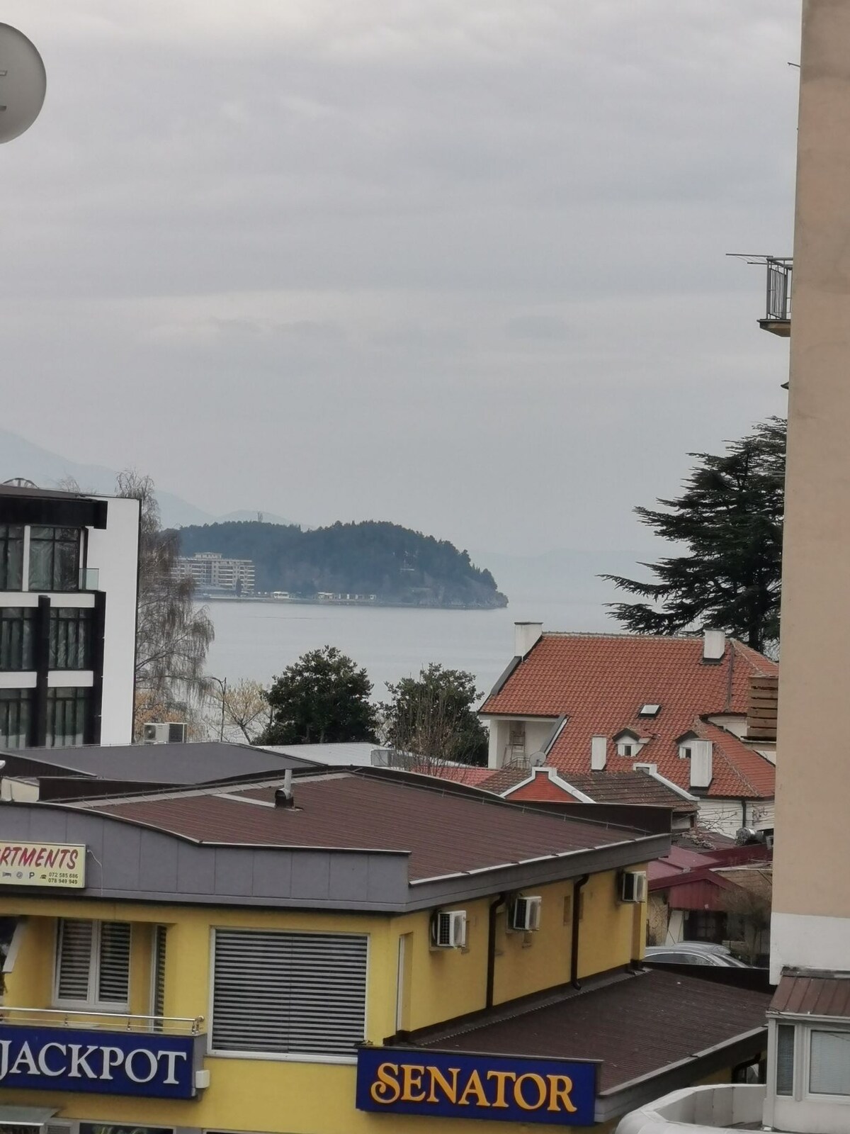 A view of the lake can be seen in the distance, framed by the rooftops of nearby buildings. Some structures display colorful signage, while trees provide a touch of greenery. The sky is overcast, casting a soft light over the scene.
