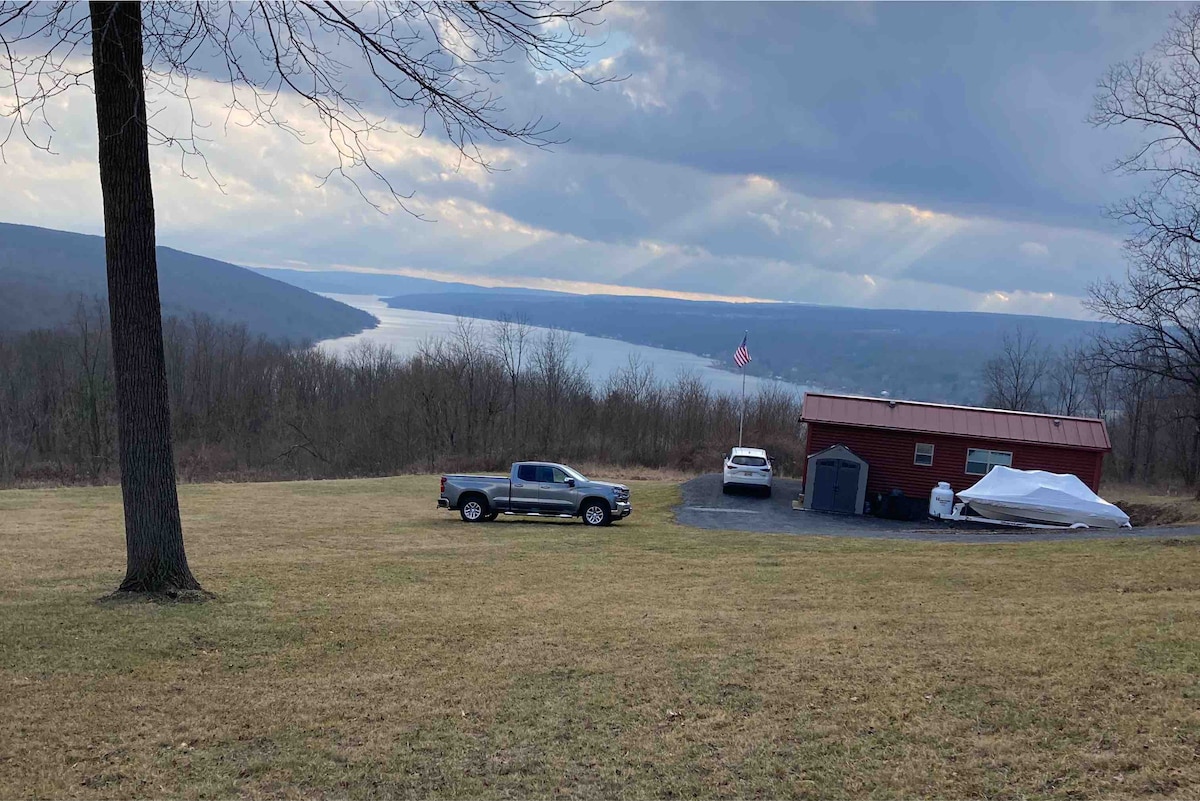 A scenic view captures the tiny house on wheels nestled within green grass, overlooking a serene body of water. A few vehicles are parked nearby, with a gentle slope leading towards the lake in the distance. Cloudy skies add a calm atmosphere to the landscape.