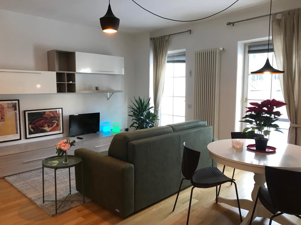 A bright living area features a comfortable green sofa positioned in front of a modern entertainment unit. A round dining table with black chairs is visible, accompanied by a decorative plant and a small flower arrangement. Natural light filters through large windows, enhancing the inviting space.