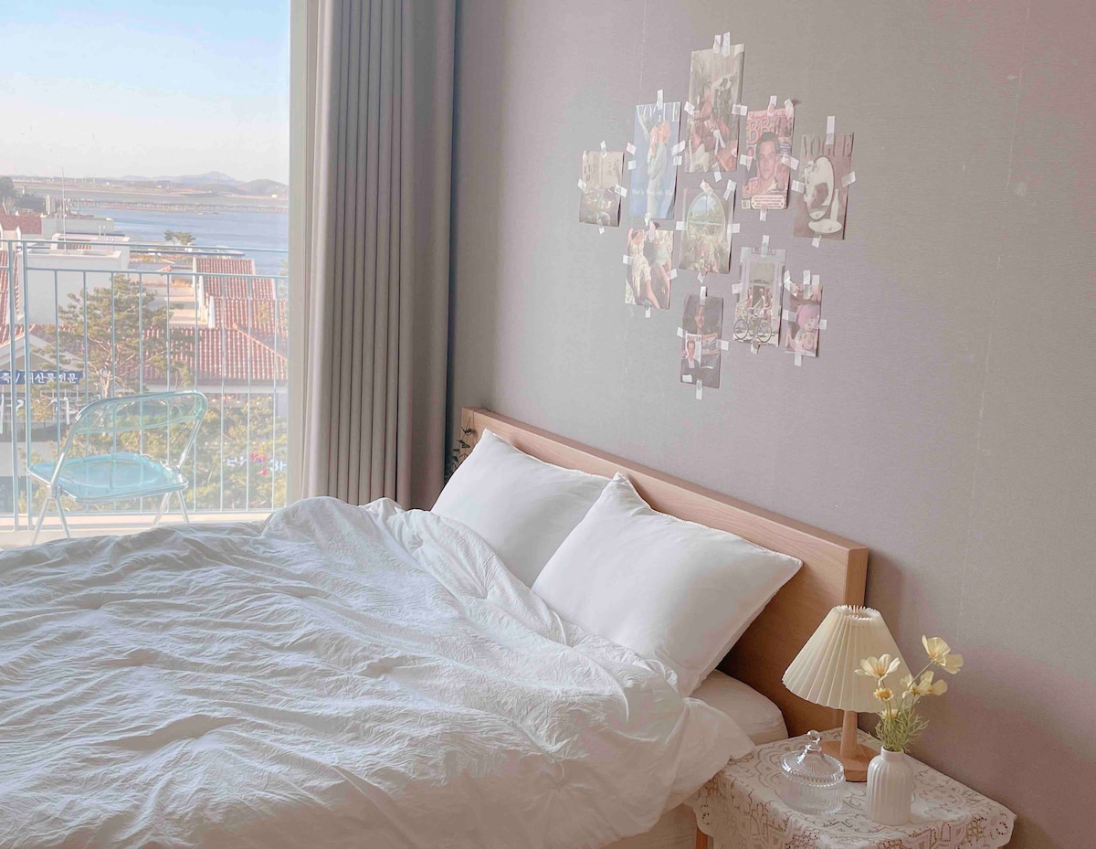 A spacious bed is dressed in soft white linens, complemented by a small bedside lamp and a floral arrangement. A wall adorned with mounted photos adds a personal touch. Natural light enters through large windows, offering a view of the sea and surrounding landscape.