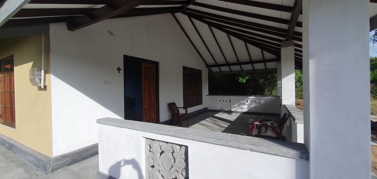 A spacious outdoor terrace is presented with a sloped roof and beams, featuring simple wooden seating for relaxation. Light pours in from large windows, and the surrounding area is enriched by lush greenery, providing a calm environment for guests to enjoy.