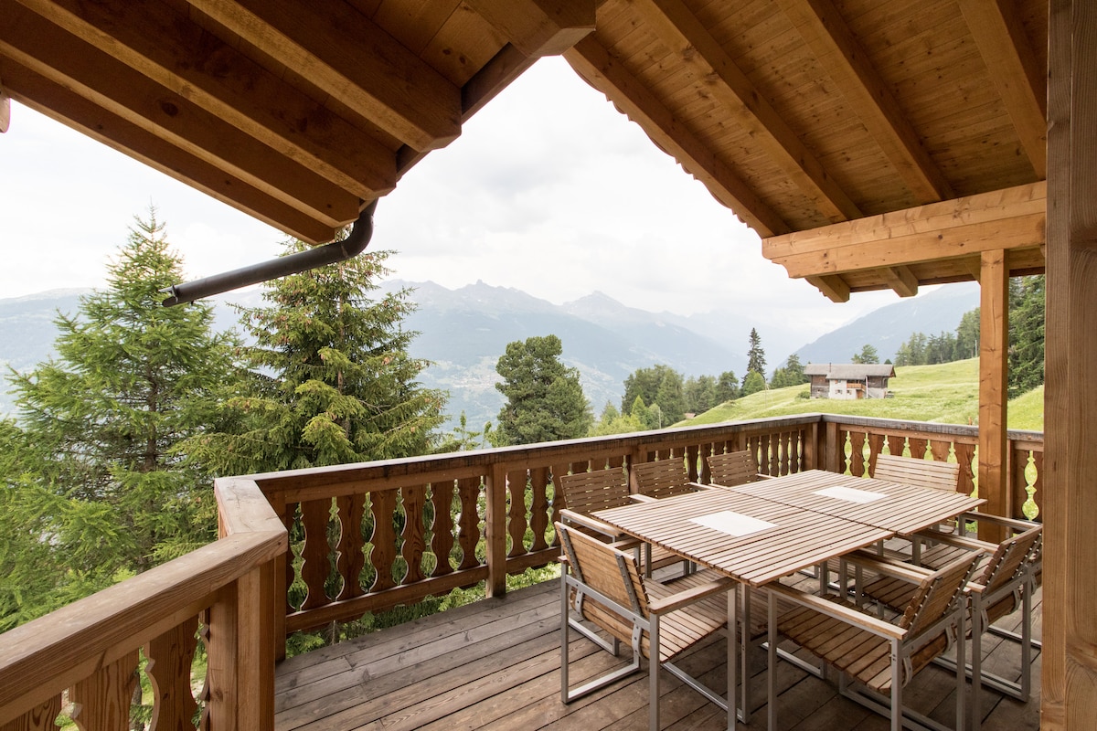 An outdoor terrace featuring a wooden table surrounded by chairs offers a serene setting for meals. The structure's traditional wooden beams provide charm, while expansive mountain views can be seen in the background, enhancing the peaceful ambiance.