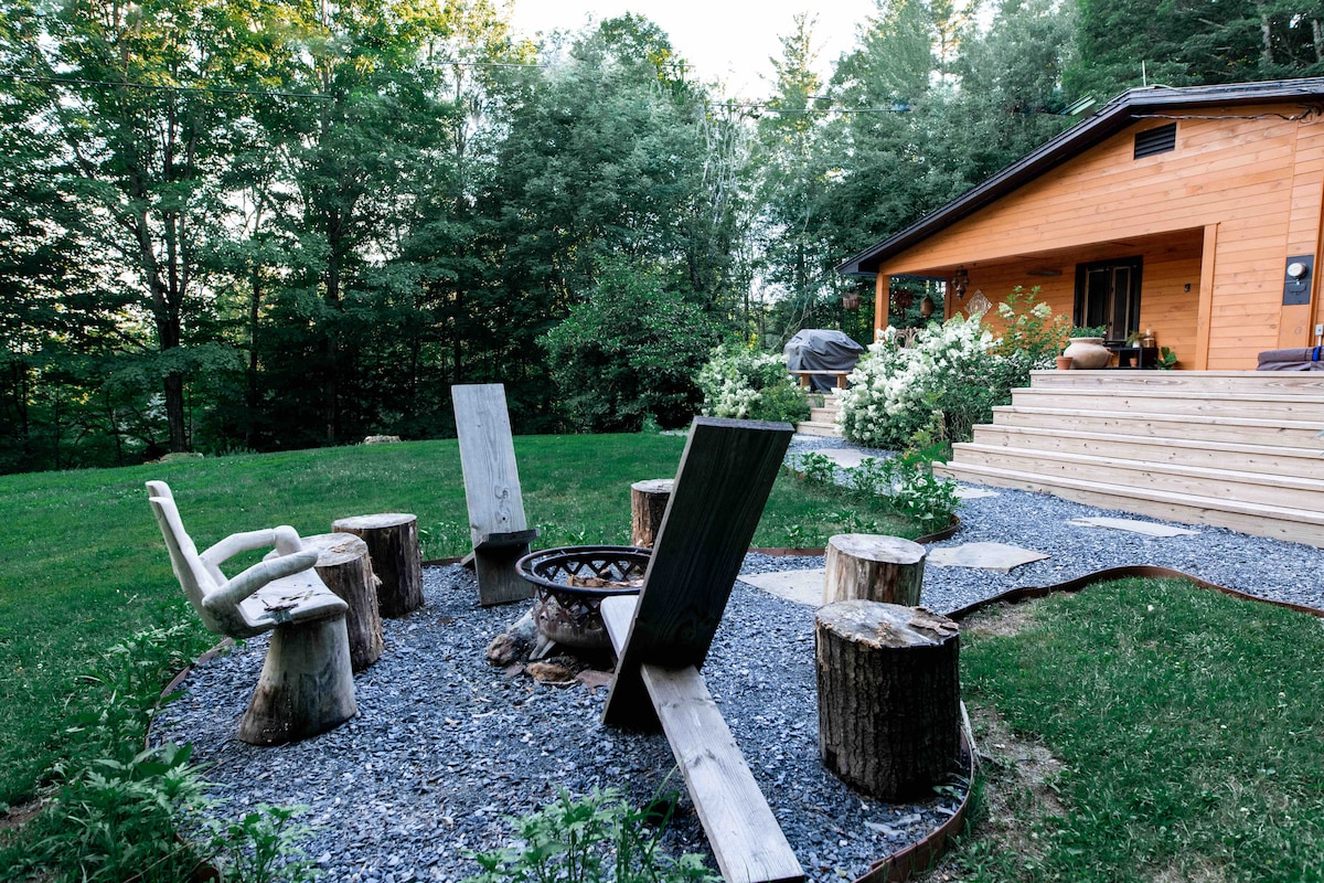 An outdoor seating area features a circular arrangement of wooden stumps and chairs surrounding a central fire pit. Lush greenery and shrubs provide a natural backdrop to the wooden structure of the house in the distance, complemented by a stone pathway leading to the entrance.