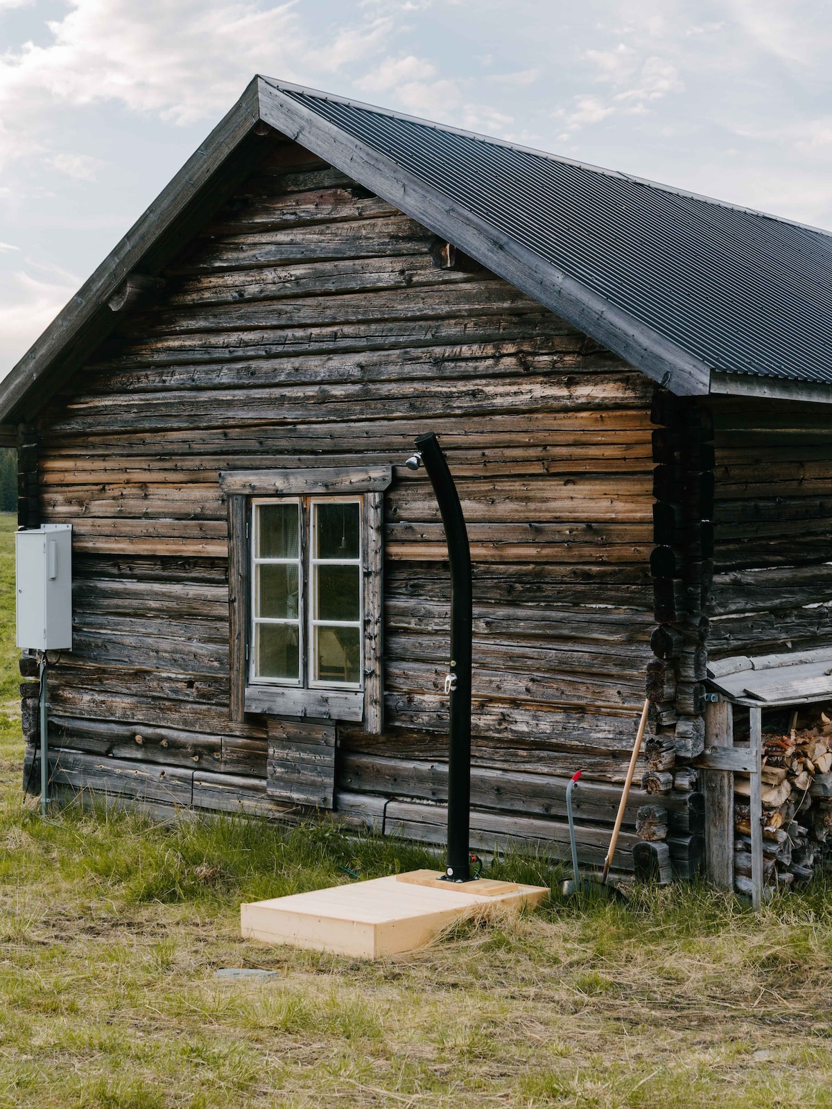 An outdoor shower is positioned against the rustic wooden exterior of the cottage. The structure features a traditional design with a metal roof and a single window. Grass surrounds the area, contributing to the natural setting.
