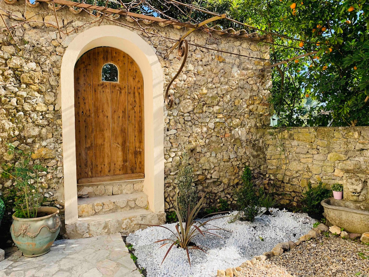 A rustic stone wall surrounds a welcoming wooden door framed by an arch. Gravel and plants create a tranquil outdoor space, with white stones accentuating the garden. Green vines drape gracefully from above, adding to the natural charm of the entrance.