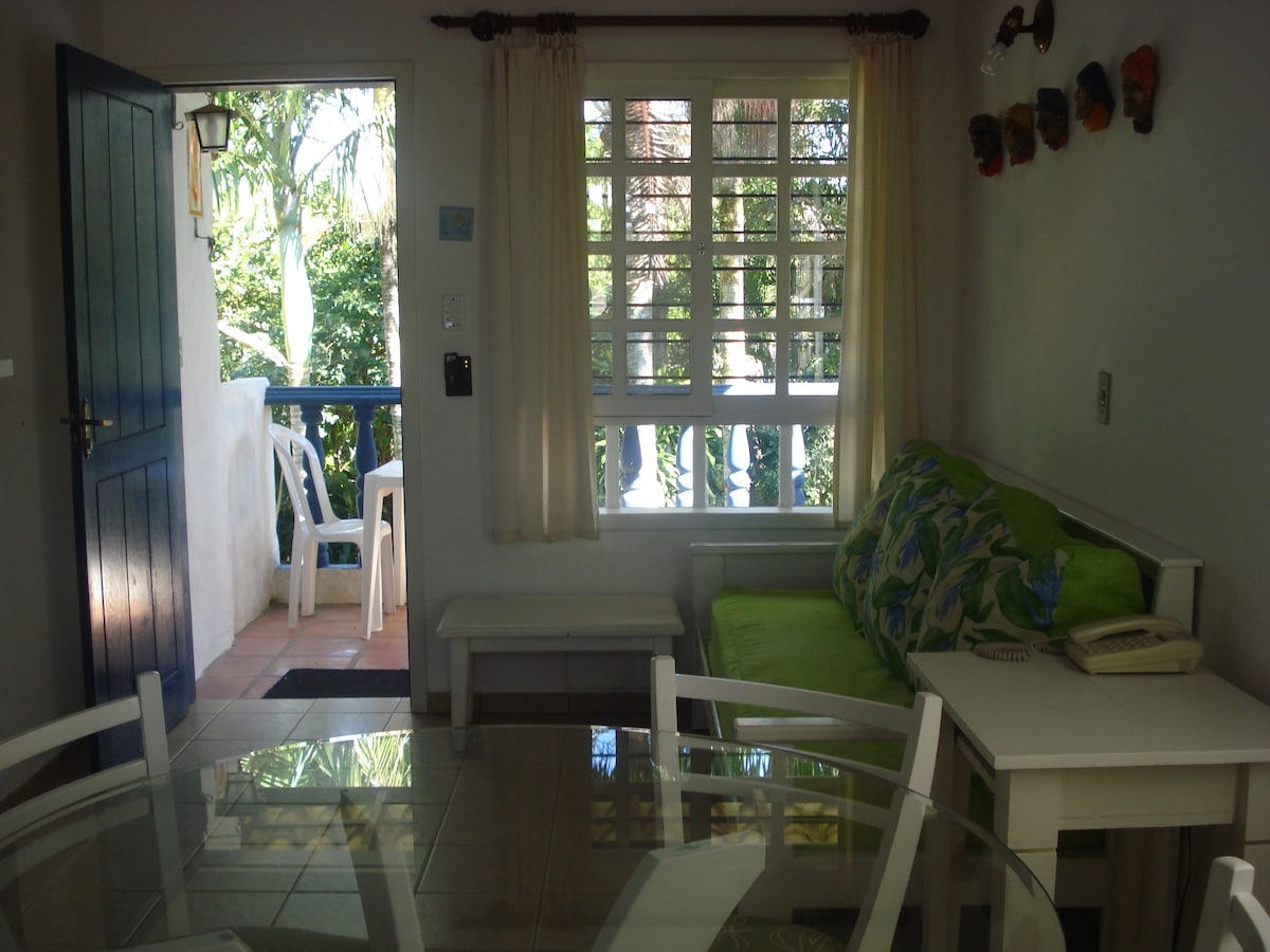 A well-lit living area features a white dining table surrounded by chairs. A green sofa bed with floral accents is placed beside a window, allowing natural light to flow in. The entrance is visible, showcasing a door leading to an outdoor patio.