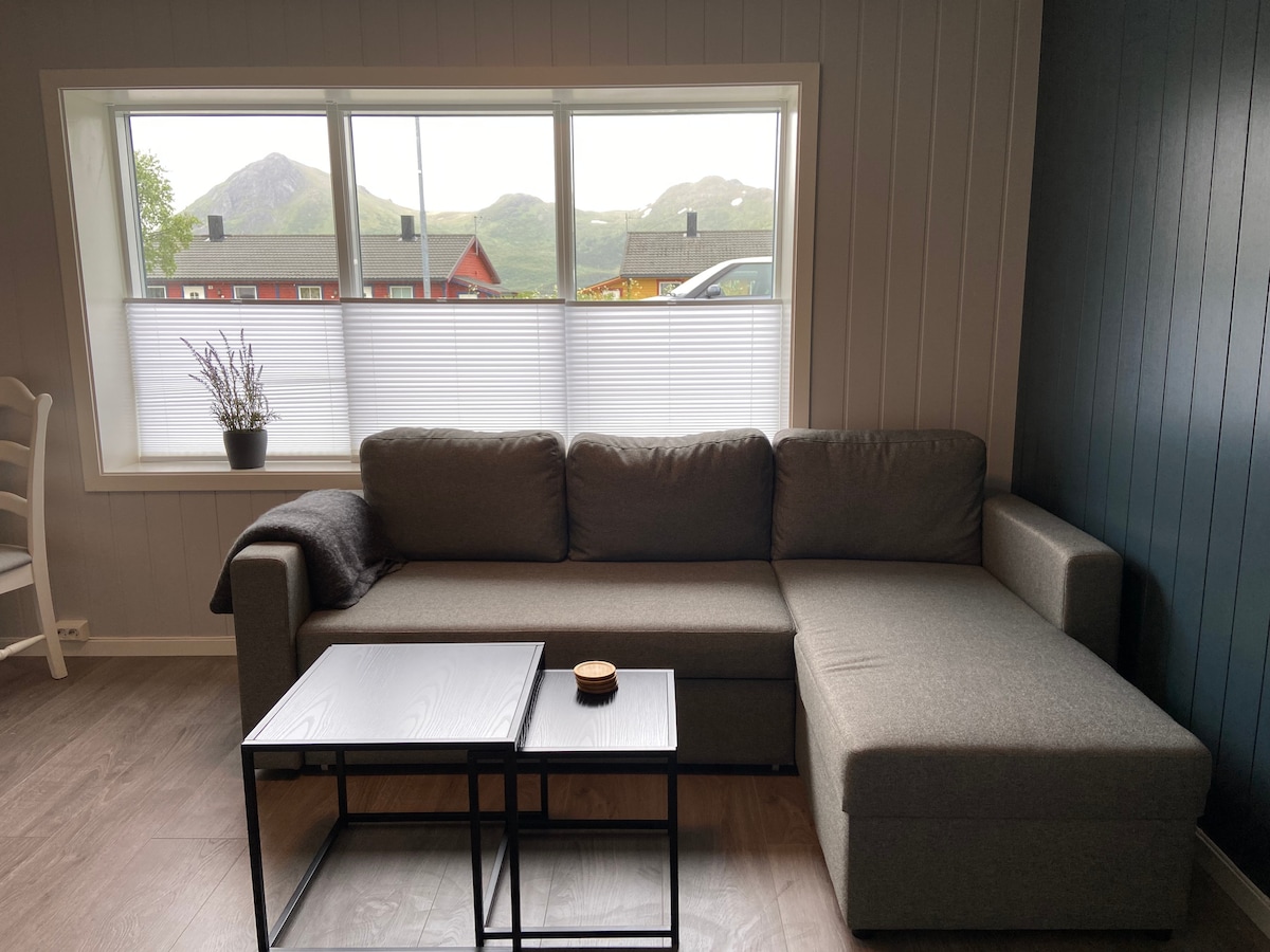 A living room corner features a gray sectional sofa positioned beside a black coffee table. Large windows allow natural light to flow in, framed by white blinds. Scenic mountain views are visible through the windows, enhancing the space's tranquility.