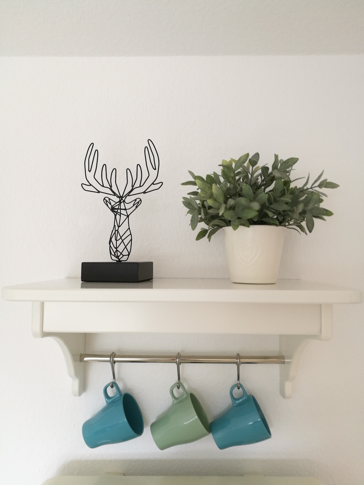 A white shelf features a decorative wire sculpture of deer antlers on a black base. A potted plant with green foliage sits beside it. Three mugs in shades of blue and green are hanging from hooks underneath the shelf, adding a functional touch.