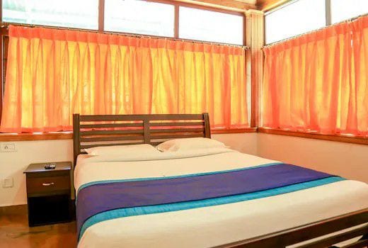 A cozy bedroom features a spacious bed with white linens and a blue accent stripe. Bright orange curtains frame the large windows, allowing natural light to fill the room. A bedside table is placed next to the bed, contributing to the functional layout.