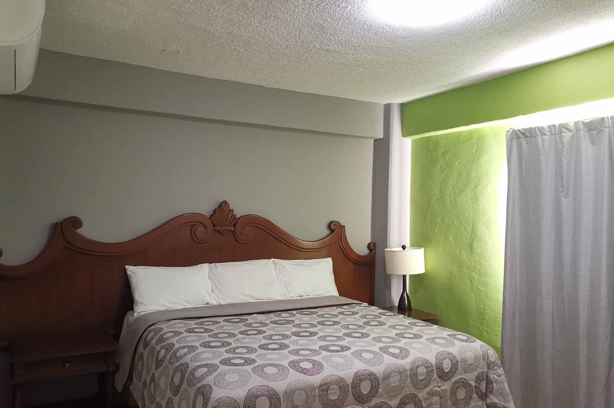 The sleeping area features a spacious bed with a patterned blanket, complemented by neutral-tone walls. A bedside table with a lamp provides soft lighting. Natural light enters through a window with sheer curtains, while a vibrant green accent wall adds a pop of color.