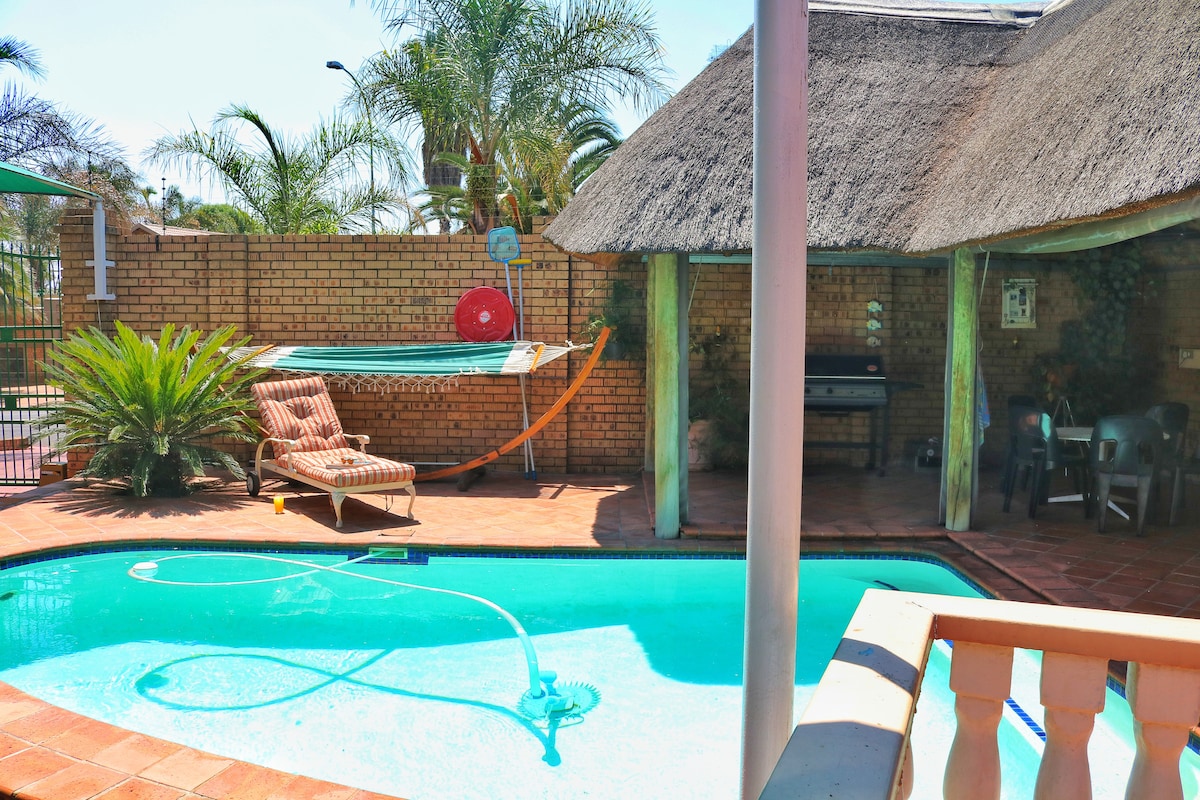 A private outdoor space features a swimmer-friendly pool surrounded by brick paving. A thatched lapa offers a seating area with chairs and a barbecue grill, while a hammock and sun loungers provide relaxation options. Nearby palm trees add to the serene ambiance.