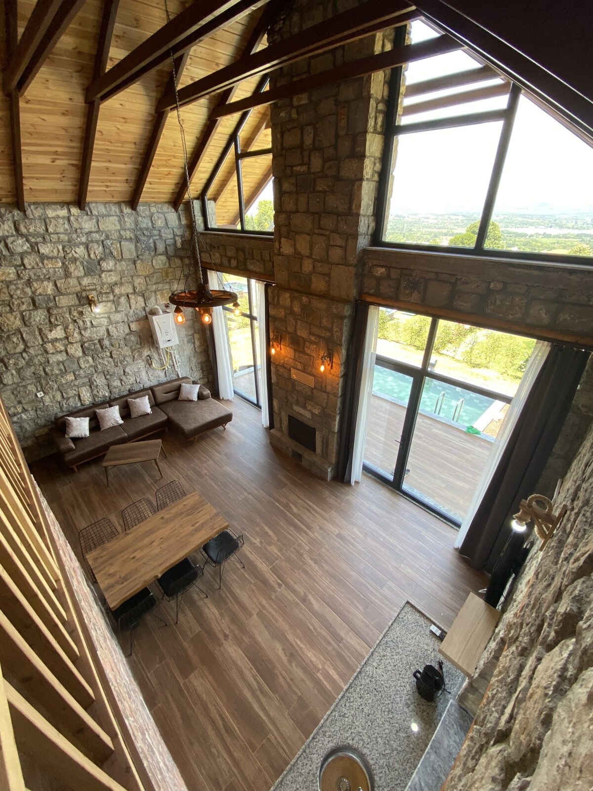 A spacious living area showcases high stone walls and a wooden ceiling, complemented by large glass doors that invite natural light. A comfortable seating arrangement is positioned near the fireplace, with a dining table visible in the foreground, providing a welcoming atmosphere.