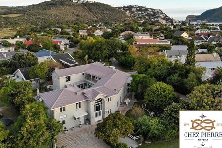 Aerial view of 'Chez Pierre' reveals a spacious two-story house surrounded by lush greenery and well-maintained gardens. The architectural design features a prominent entrance with large windows. Nearby neighborhoods and hills form a scenic backdrop, highlighting the serene atmosphere of the location.