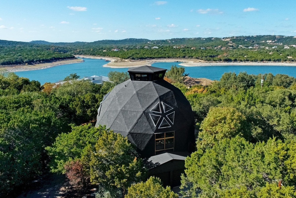 The Geodome is presented from an aerial perspective, showcasing its unique geodesic structure. Surrounded by lush greenery, the dome contrasts with the serene blue waters of the nearby lake. Treetops frame the view, enhancing the sense of privacy and tranquility offered by this natural setting.