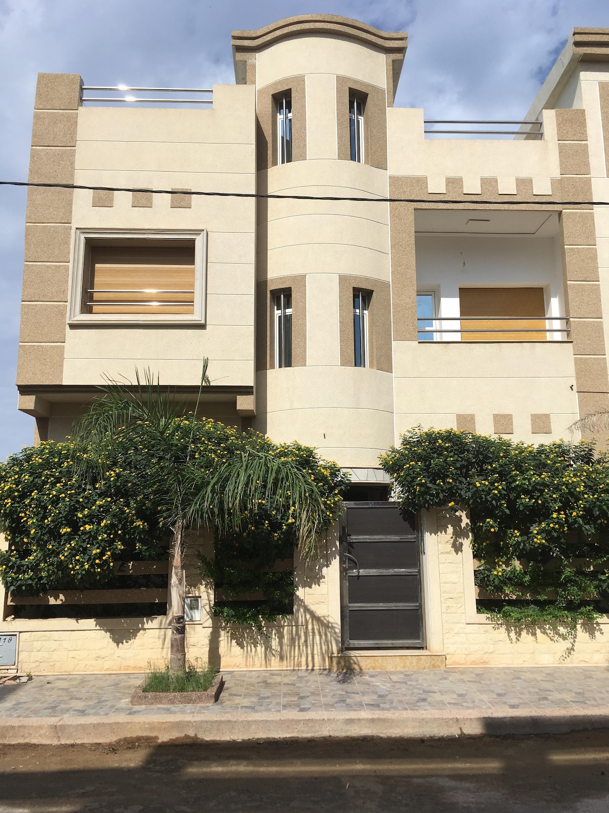 The exterior of a modern two-story building is showcased, featuring a combination of light and dark stone textures. Large windows provide natural light, while greenery decorates the façade. A wrought iron gate leads to the entrance, surrounded by well-maintained landscaping.