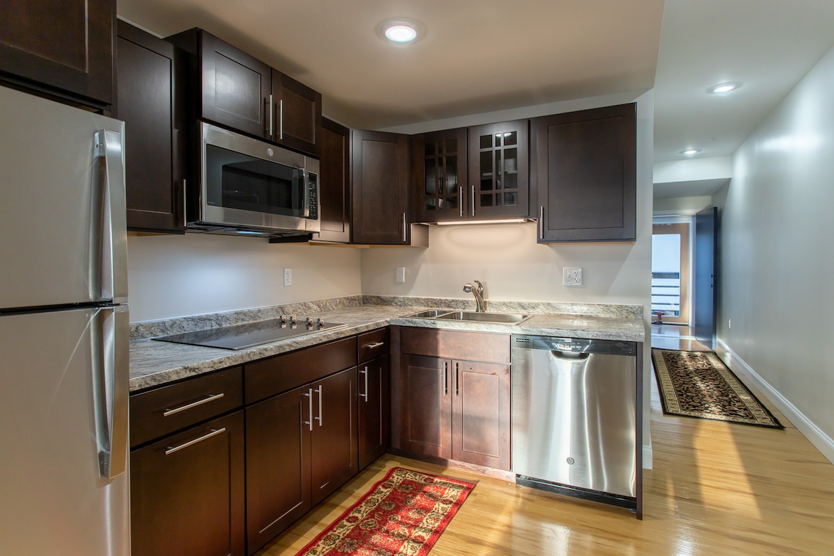 A well-appointed kitchen features dark cabinetry complemented by granite countertops. Stainless steel appliances, including a refrigerator, microwave, and dishwasher, are visible. A decorative rug adds a touch of color, while ample lighting enhances the clean lines of the space.