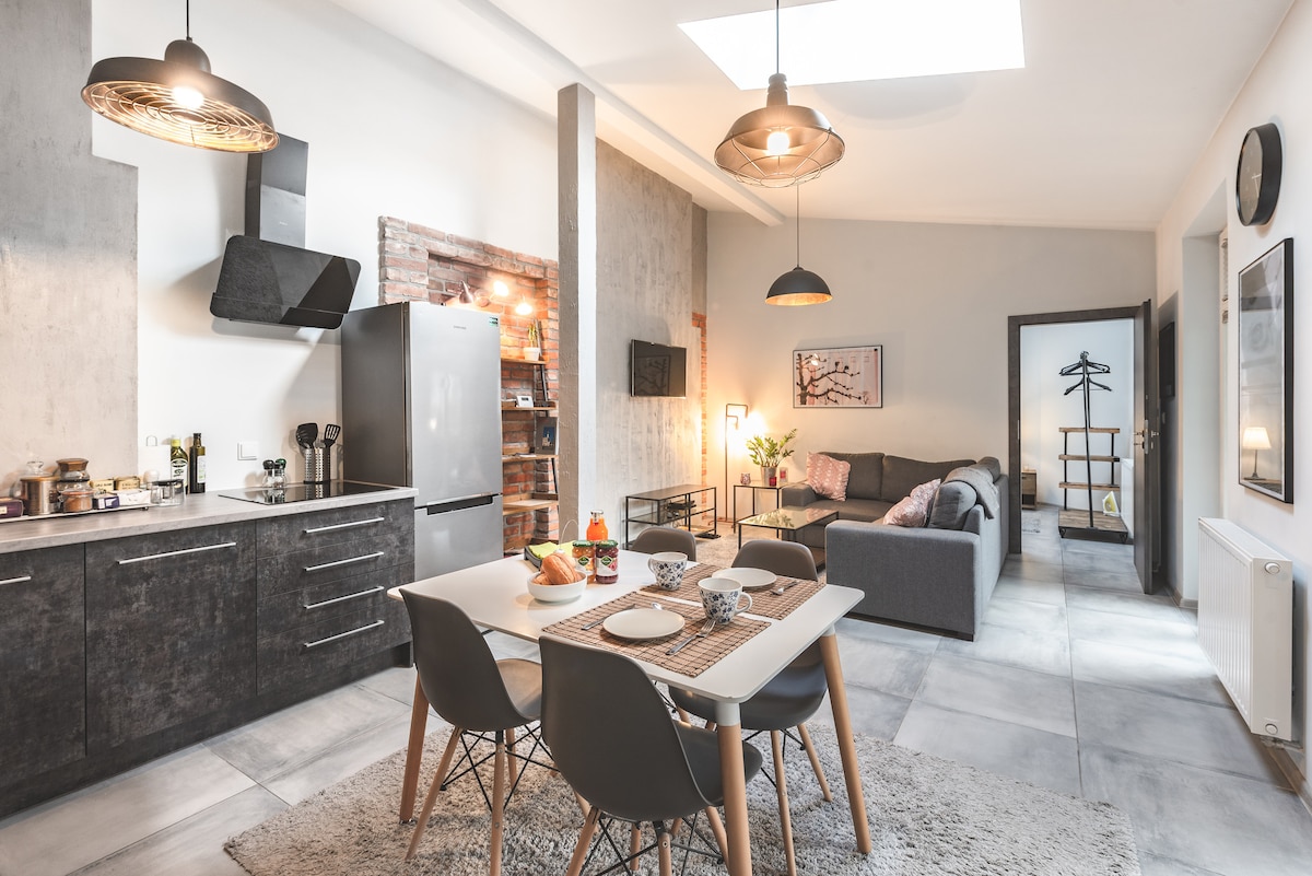 An open kitchen area featuring modern appliances, including a refrigerator and stove, is complemented by a dining table with four chairs. A cozy living room space with a gray sofa is visible, illuminated by skylights and pendant lights that enhance the airy atmosphere.