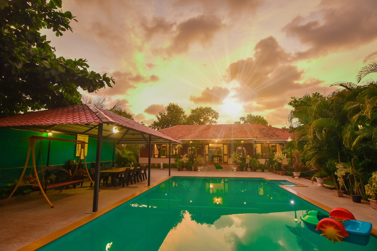 The image captures a serene villa view as the sun sets behind lush greenery. A large pool reflects the warm colors of the sky, while a covered seating area is visible alongside the pool, offering a comfortable space for relaxation surrounded by nature.