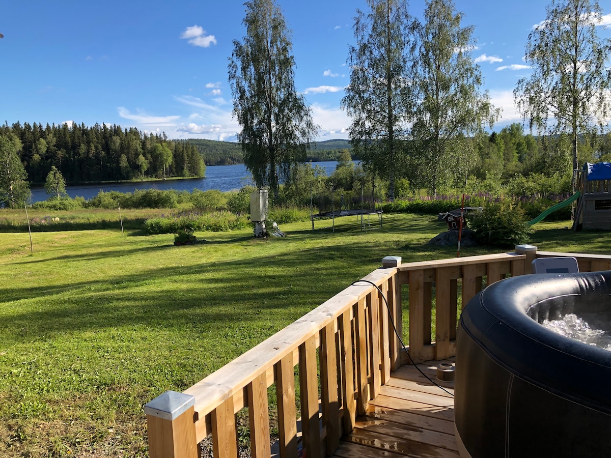 A deck overlooks a spacious grassy area leading to a serene lake, surrounded by trees. A hot tub is positioned nearby, inviting relaxation. The sky features scattered clouds, creating a calming atmosphere, while the lush greenery enhances the natural setting.
