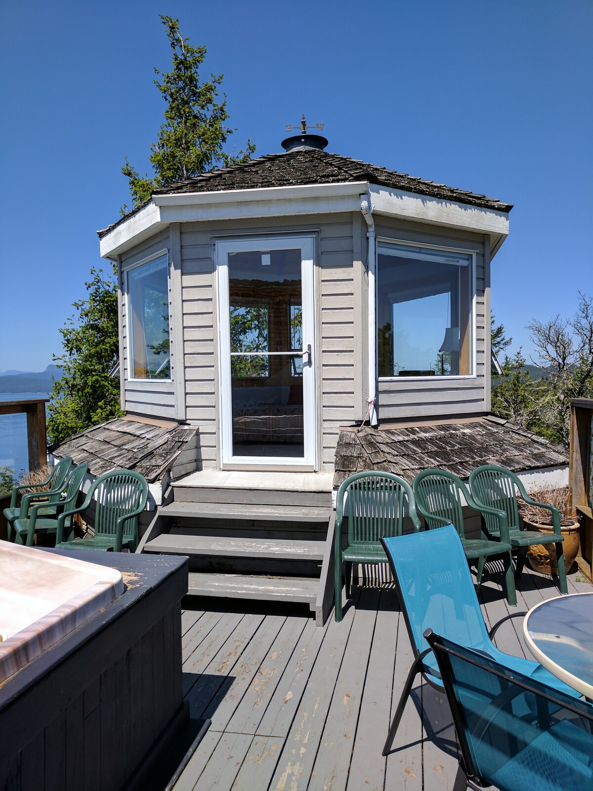 A circular tower structure rises, featuring large windows that provide panoramic views. Steps lead up to the entrance, adjacent to a seating area with multiple chairs. A hot tub rests nearby, complemented by a deck that invites relaxation in a peaceful outdoor setting.