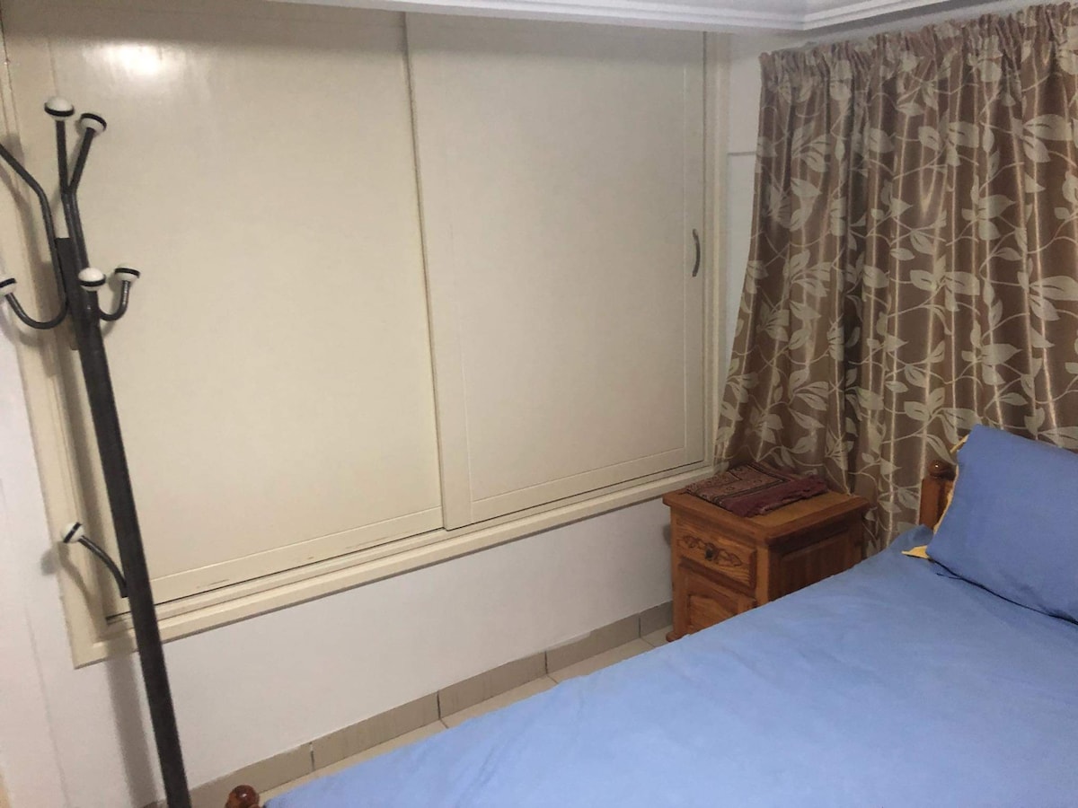 A cozy sleeping area features a spacious bed topped with a blue blanket. A small wooden nightstand is positioned beside the bed, while a coat rack stands against the wall. Natural light filters softly through a window with light-colored curtains.