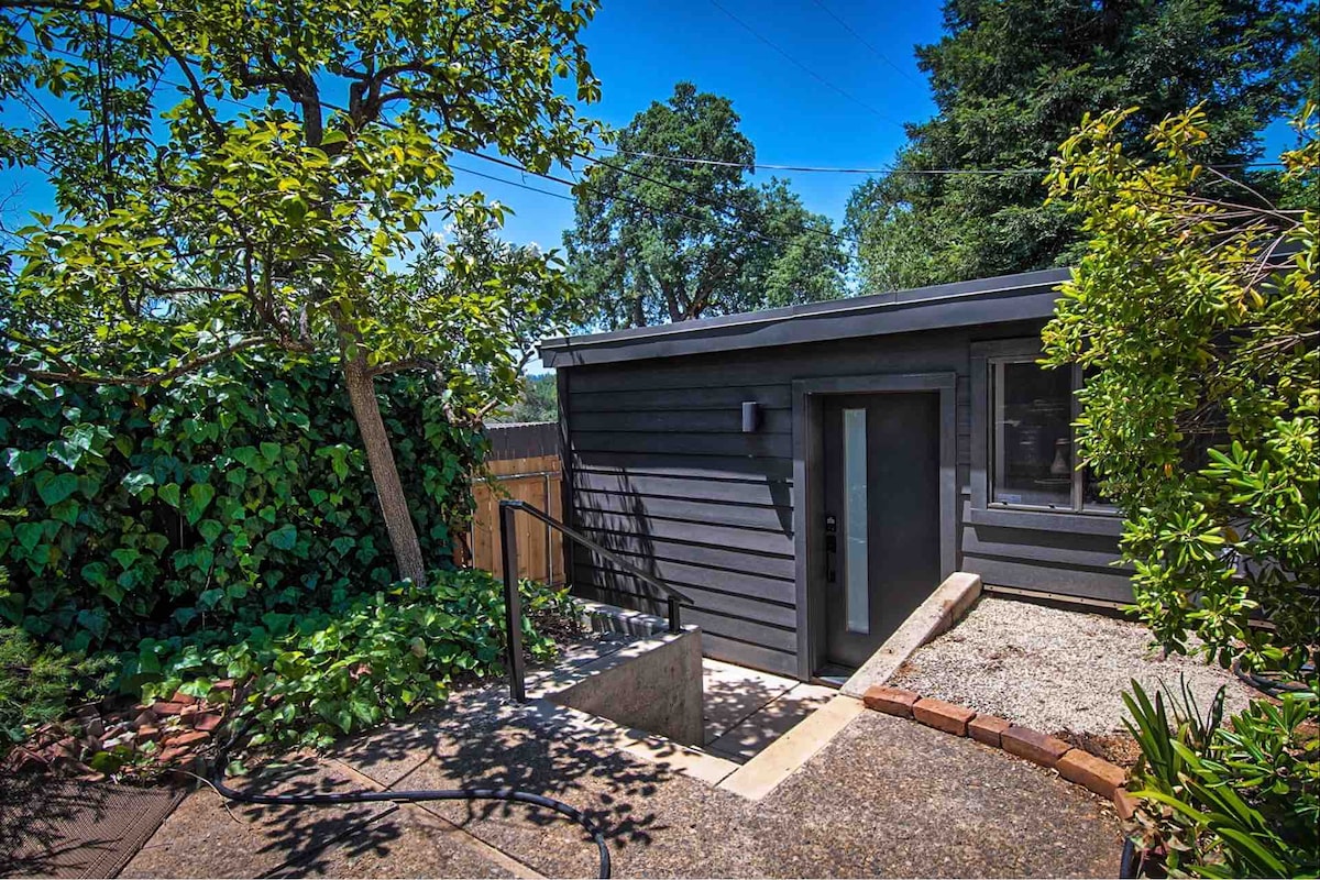 An exterior view of the detached one-bedroom unit is shown, featuring a simple, modern design with charcoal-colored siding. The entrance is accessible by a few steps, surrounded by lush greenery, including a tree and climbing vines, contributing to a serene, natural setting.