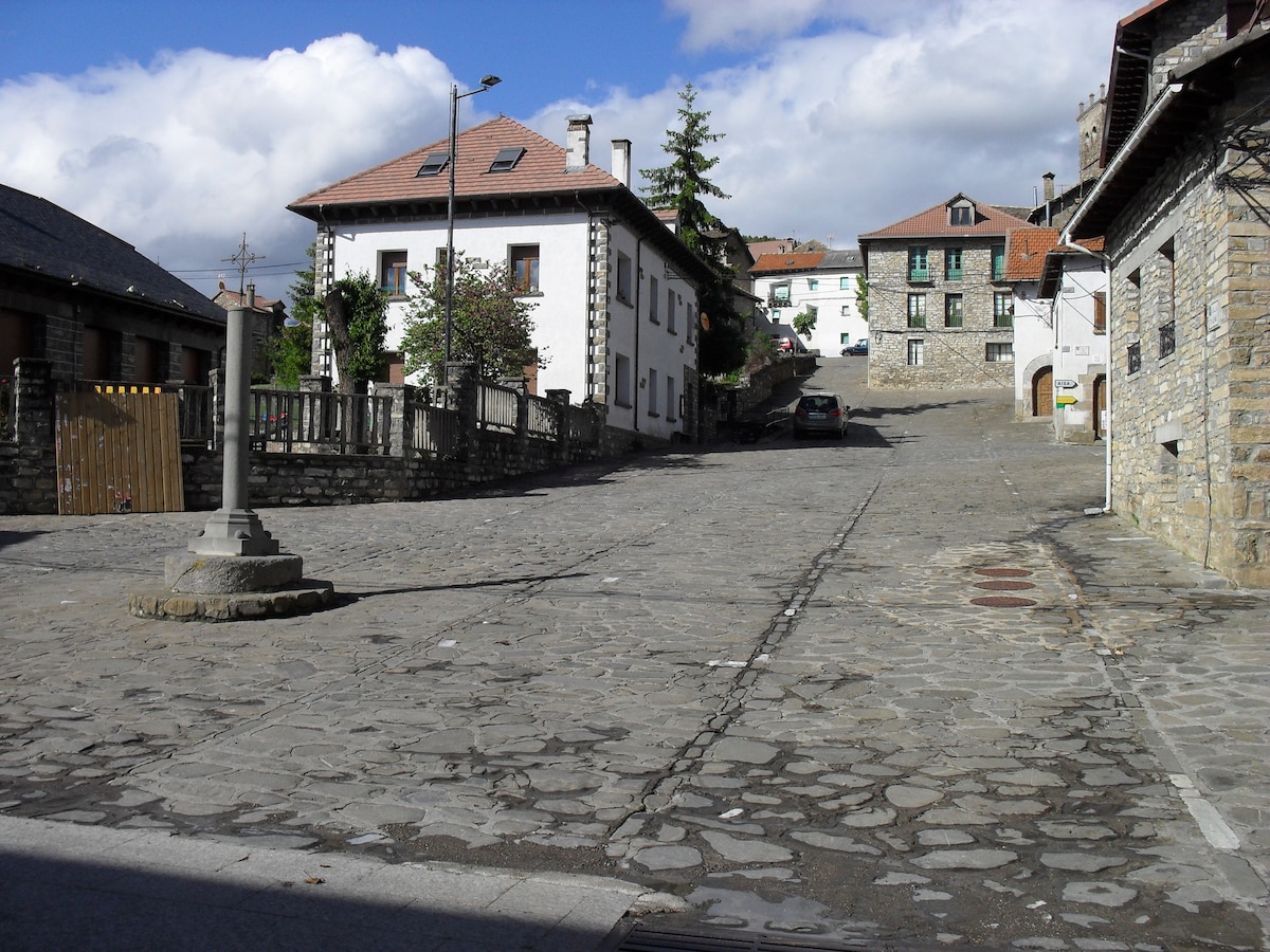 A tranquil street scene is depicted, showcasing a cobblestone road lined with charming buildings. The structures feature a mix of stone and traditional architecture. Lush greenery is present, with trees and shrubs dotting the landscape, all under a partly cloudy sky.