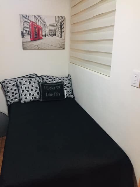 The sleeping area features a compact bed covered in a solid black bedspread, accented by patterned pillows. A wall art piece depicting a London telephone booth is displayed above the bed, while textured wall panels add visual interest to the room.