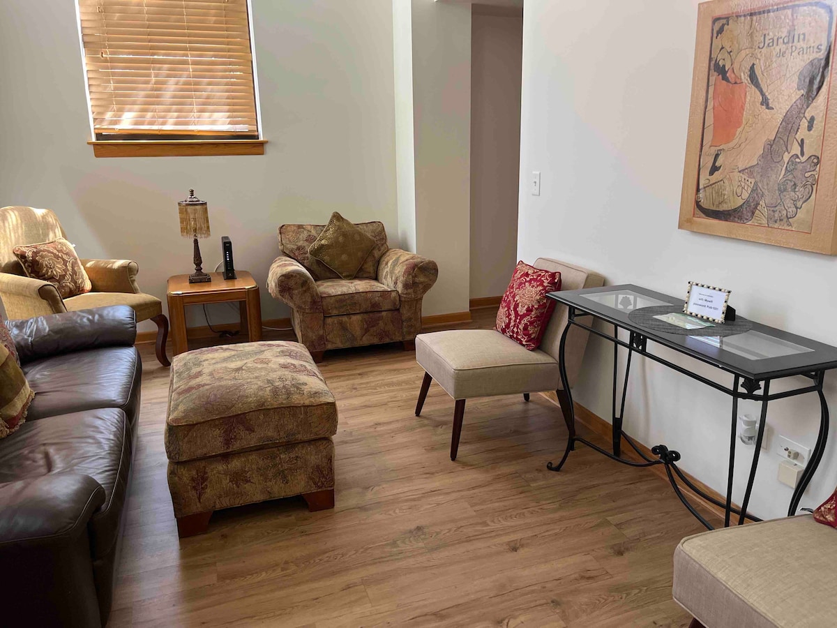 A relaxed living area is presented with a combination of plush seating, including a dark leather sofa and floral-patterned chairs. A small, decorative table with a lamp adds warmth, while a vintage-style wall art piece enhances the decor. Natural light filters through partially closed blinds.