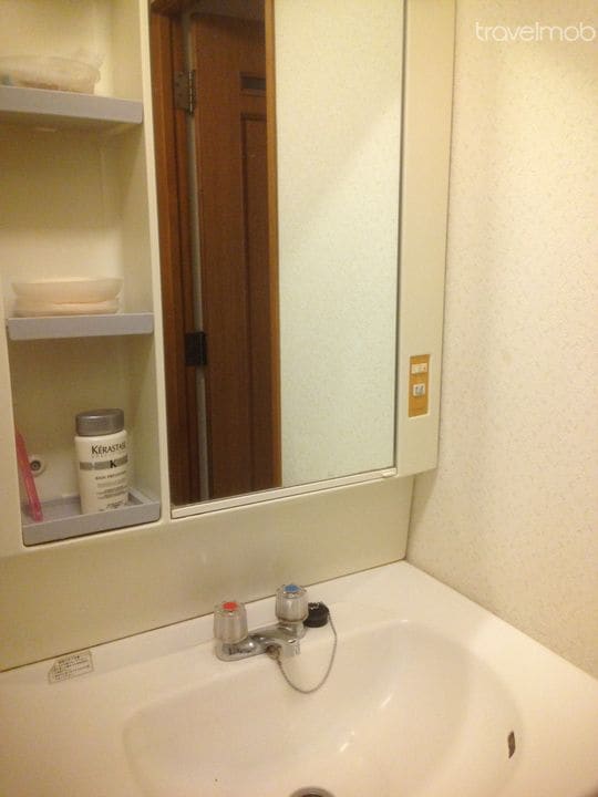 A compact bathroom area is displayed, featuring a clean, white sink with a single faucet. A wall-mounted mirror is positioned above the sink, reflecting a portion of the room. Shelves are installed beside the sink, containing personal care items in neutral containers.