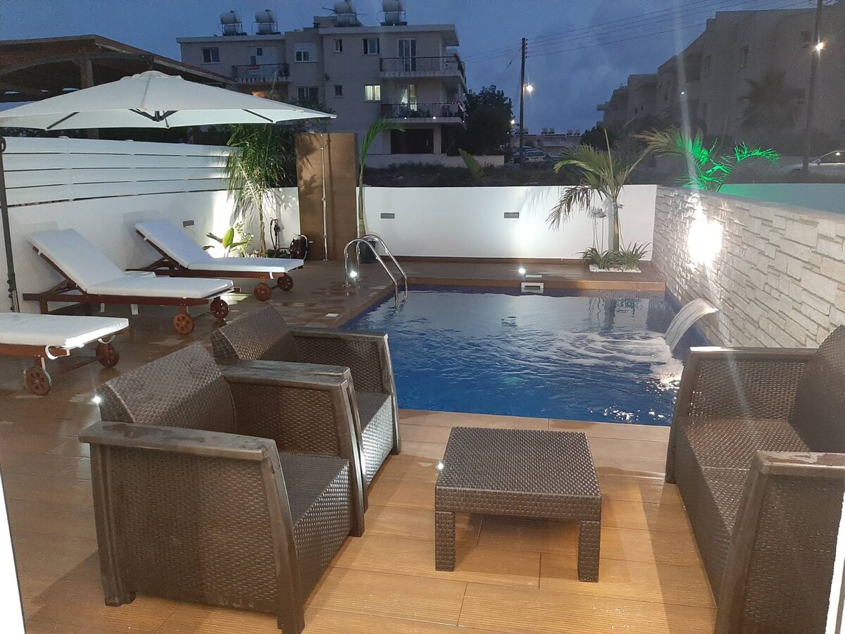 A private swimming pool is surrounded by a wooden deck and accented with lounge chairs. A small table is positioned nearby. Soft lighting casts a calming glow on the water, while palm plants add a touch of greenery to the space.