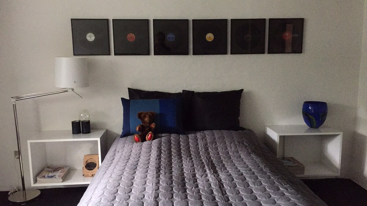 A cozy sleeping area features a spacious bed with a textured gray quilt. Two bedside tables hold books and decorative items. Above the bed, framed vinyl records add a touch of personality, while a blue glass bowl and a table lamp complete the serene space.