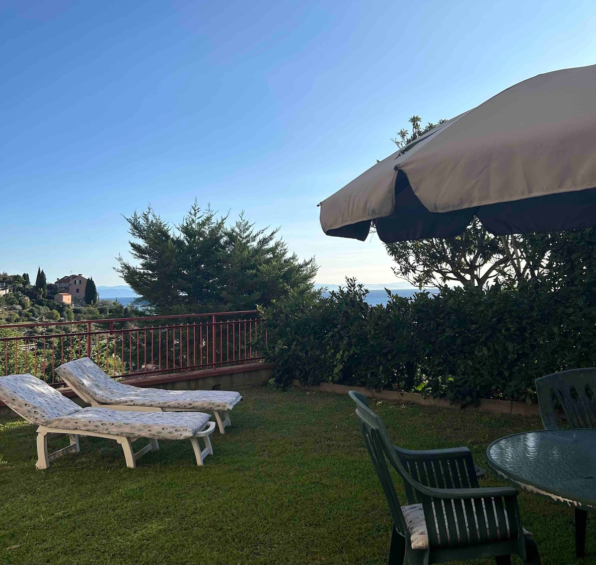 A green garden area features two lounge chairs positioned for relaxation, complemented by a small table and chairs under a large umbrella. The landscape presents gentle shrubbery and trees, with a distant view of the sea visible beyond the garden's edge.