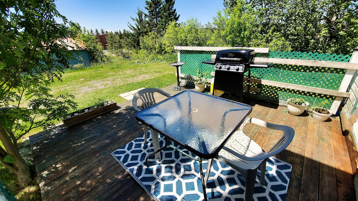 An outdoor deck features a glass-top table surrounded by two comfortable chairs. A BBQ is positioned nearby, complemented by greenery in the background. The space is adorned with a geometric rug, providing an inviting atmosphere for outdoor meals or relaxation.
