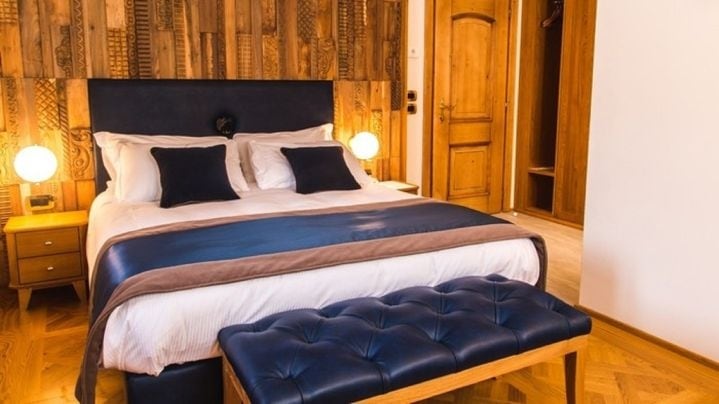 A spacious bed with a dark blue and neutral bedding arrangement is positioned against a wooden accent wall. Light fixtures are placed on either side of the bed. A wooden bench adds seating, while a closet area is visible in the background.