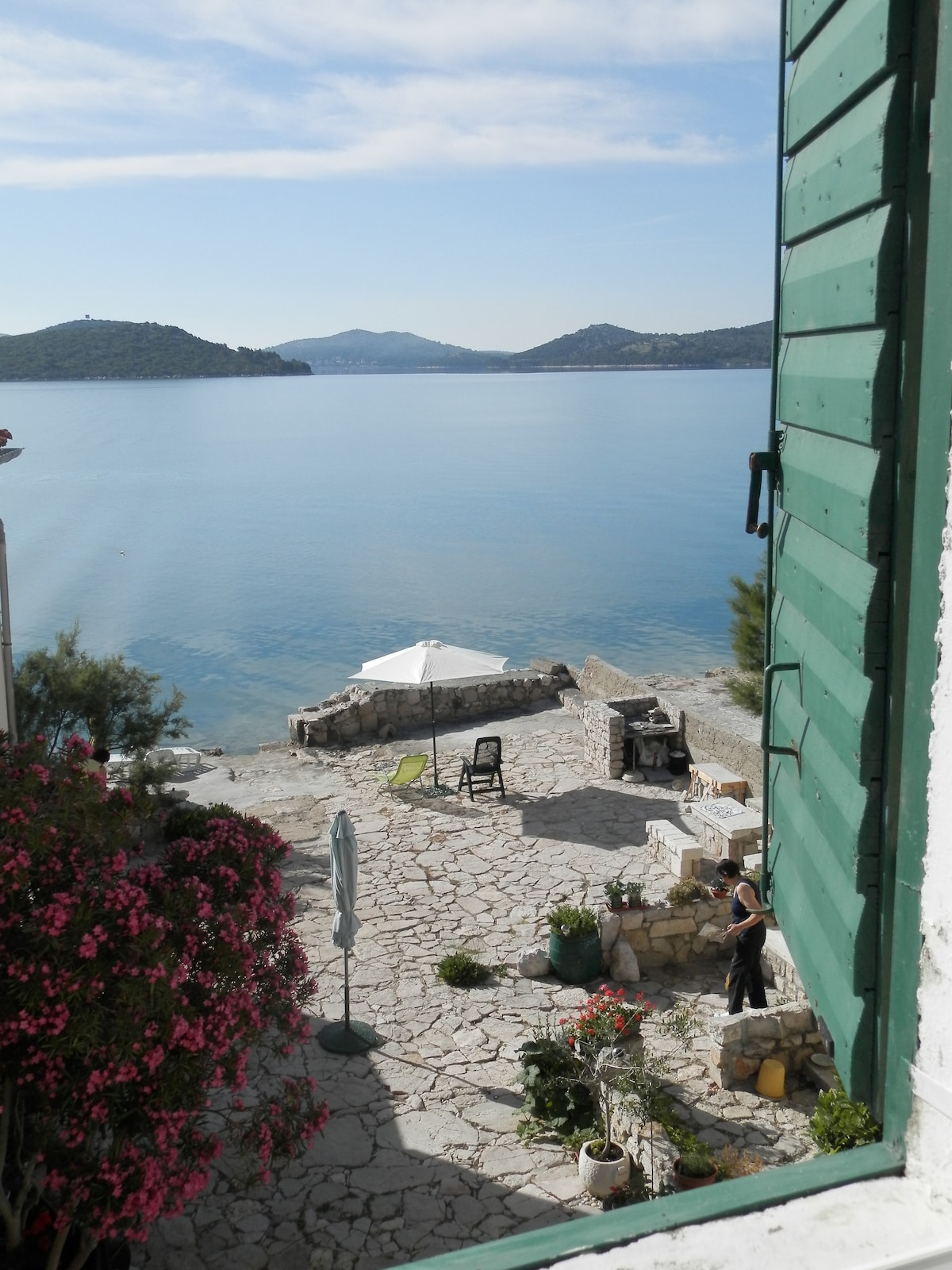 A view from an open window reveals a serene stone terrace leading to the calm sea. A sunshade and a few chairs are present, alongside a barbecue area. Vibrant flowers add color to the scene, against the backdrop of tranquil waters and distant hills.