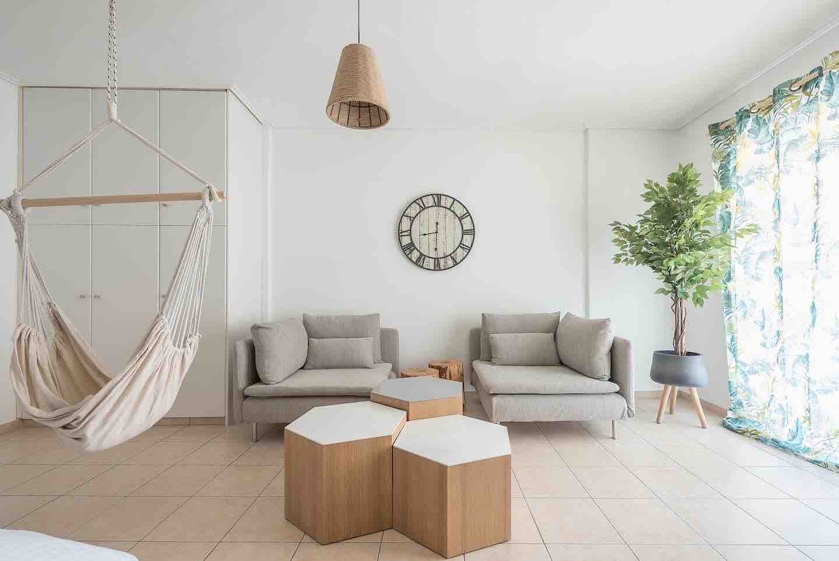 The living space features two soft grey sofas positioned across from a unique set of hexagonal wooden coffee tables. A hammock hangs from the ceiling, and a decorative wall clock adds interest. Bright, patterned curtains frame a leafy indoor plant near the window.