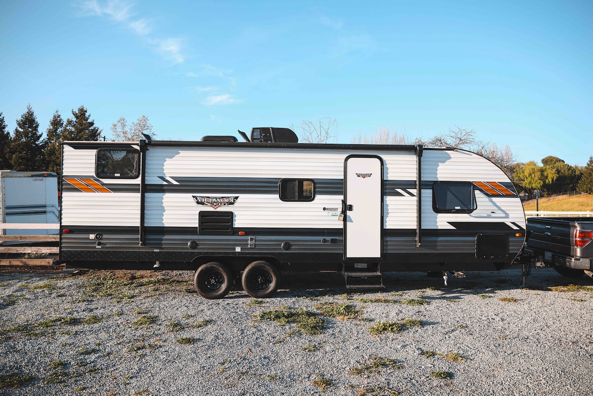 A modern travel trailer displays a sleek exterior with contrasting black and white panels, accented by bright orange stripes. The entryway is clearly visible, providing easy access. The surrounding gravel area and trees in the background contribute to a natural setting.