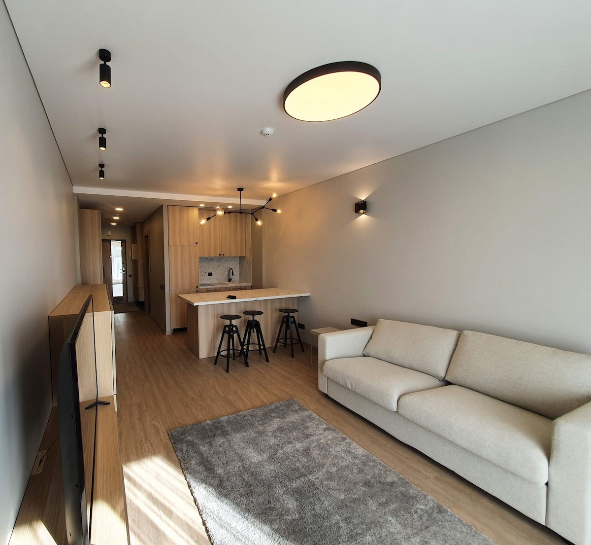 A modern living area features a soft grey color palette with warm wood accents. A spacious sectional sofa faces a low TV console. The kitchen bar, equipped with three high stools, is visible in the background, illuminated by pendant lighting.