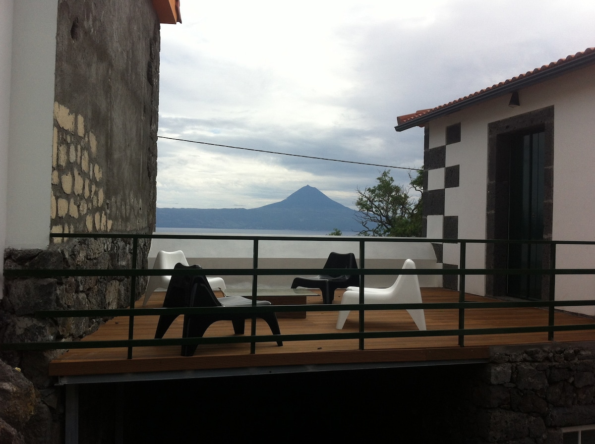 A spacious deck is depicted, featuring several modern black and white chairs arranged for leisure. In the background, a stunning view of the nearby Pico Island is visible, framed by the serene sky and surrounding greenery.