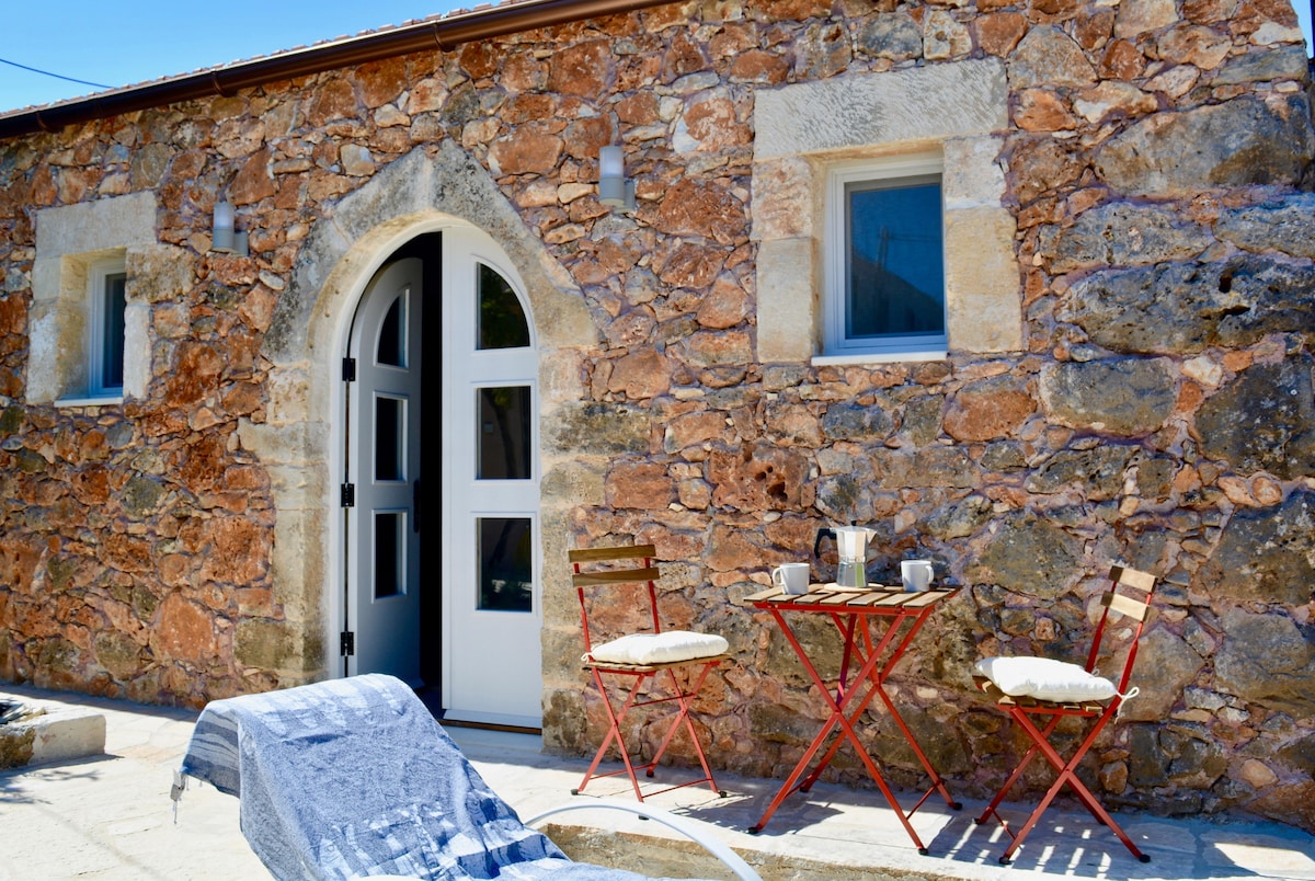 The exterior showcases a rustic stone wall with a charming archway entrance. A small bistro table with two red chairs sits invitingly outside, complemented by a sun lounger. Sunlight highlights the textured stone, creating a warm and welcoming space.
