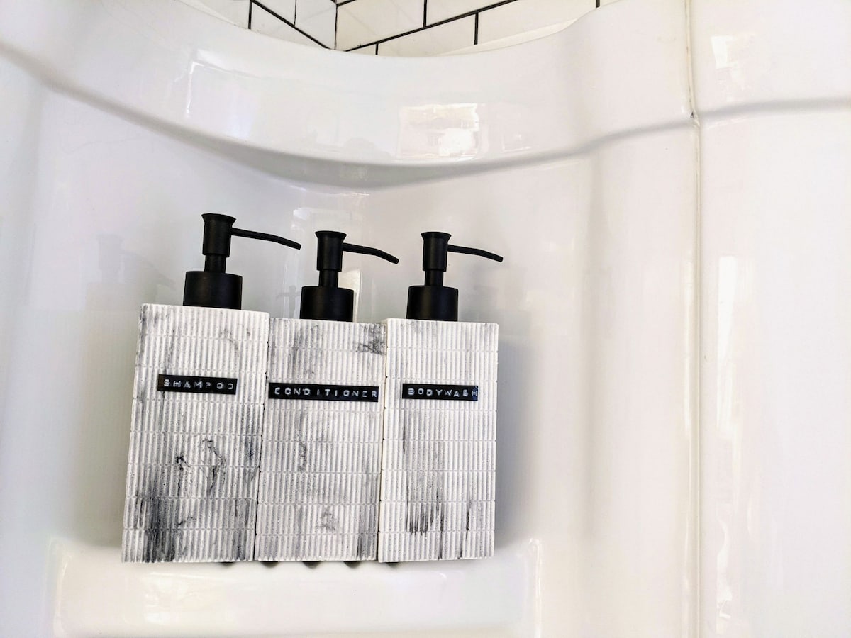 Three neatly arranged pump bottles containing shampoo, conditioner, and body wash are displayed in a shower nook. The bottles have a textured design and are labeled clearly, complementing the minimalist aesthetic of the surrounding tiled wall.