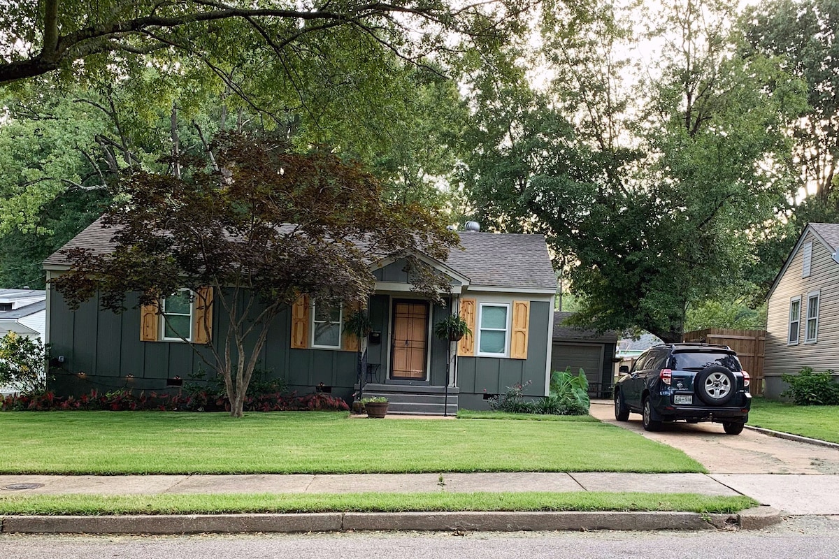 The exterior of the home features a well-maintained yard with lush green grass and flower beds. The house is painted in a muted green tone, complemented by wooden shutters. A black vehicle is parked in the driveway, with trees providing shade in the background.