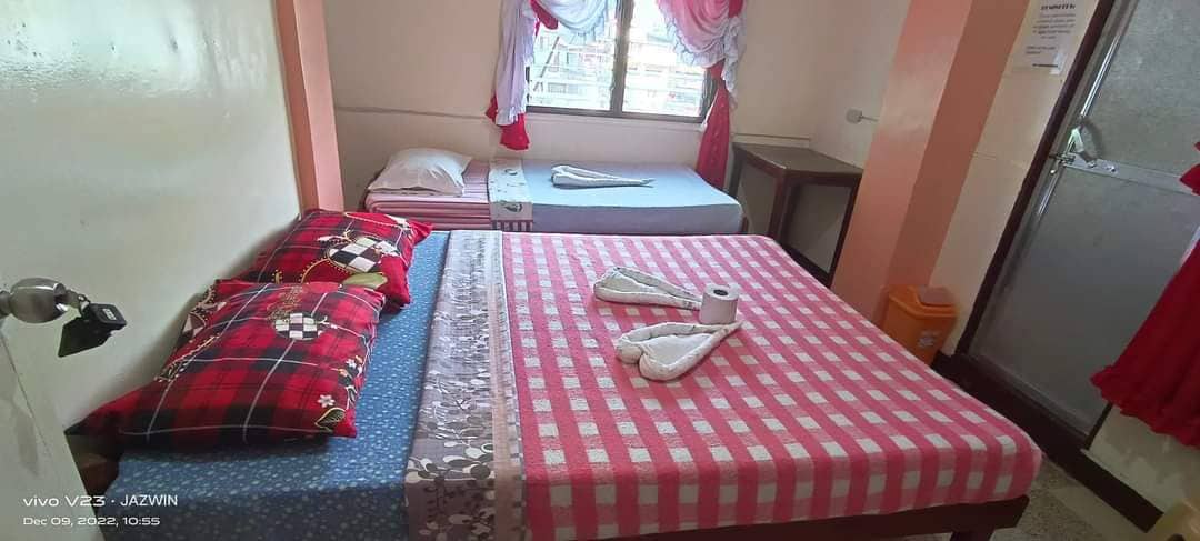 The cozy room features two beds covered with patterned linens and decorative pillows. A small table is positioned nearby, and natural light enters through a curtained window. Towels are neatly arranged on the beds, contributing to the room's tidy appearance.