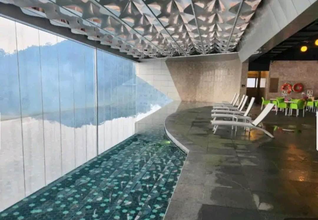 An indoor heated pool features a modern design with geometric patterns on the ceiling. Lounge chairs are positioned along the poolside, offering a place to relax. Large windows provide views of the surrounding mountains, contributing to a serene atmosphere.