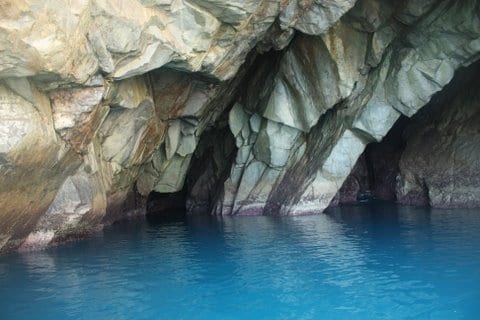 Rugged rock formations create a natural cave structure, with textured surfaces reflecting light. The entrance is framed by sharp-edged cliffs, leading to tranquil blue waters that fill the cave's interior. The calmness of the water contrasts with the solid, earthy tones of the surrounding rocks.