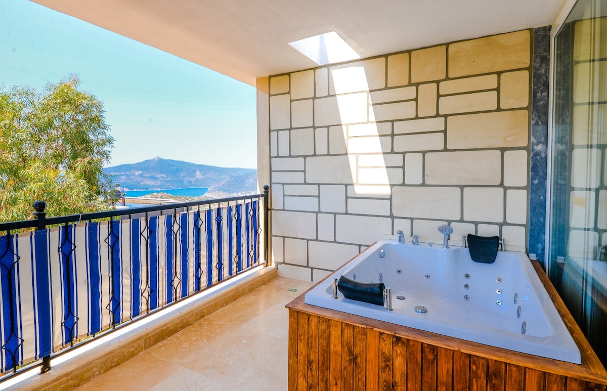 An outdoor terrace features a private jacuzzi, surrounded by wooden paneling. Light filters through the overhead opening, highlighting the spacious area. A scenic view of the hills and surrounding landscape is visible beyond the balcony railing adorned with blue accents.