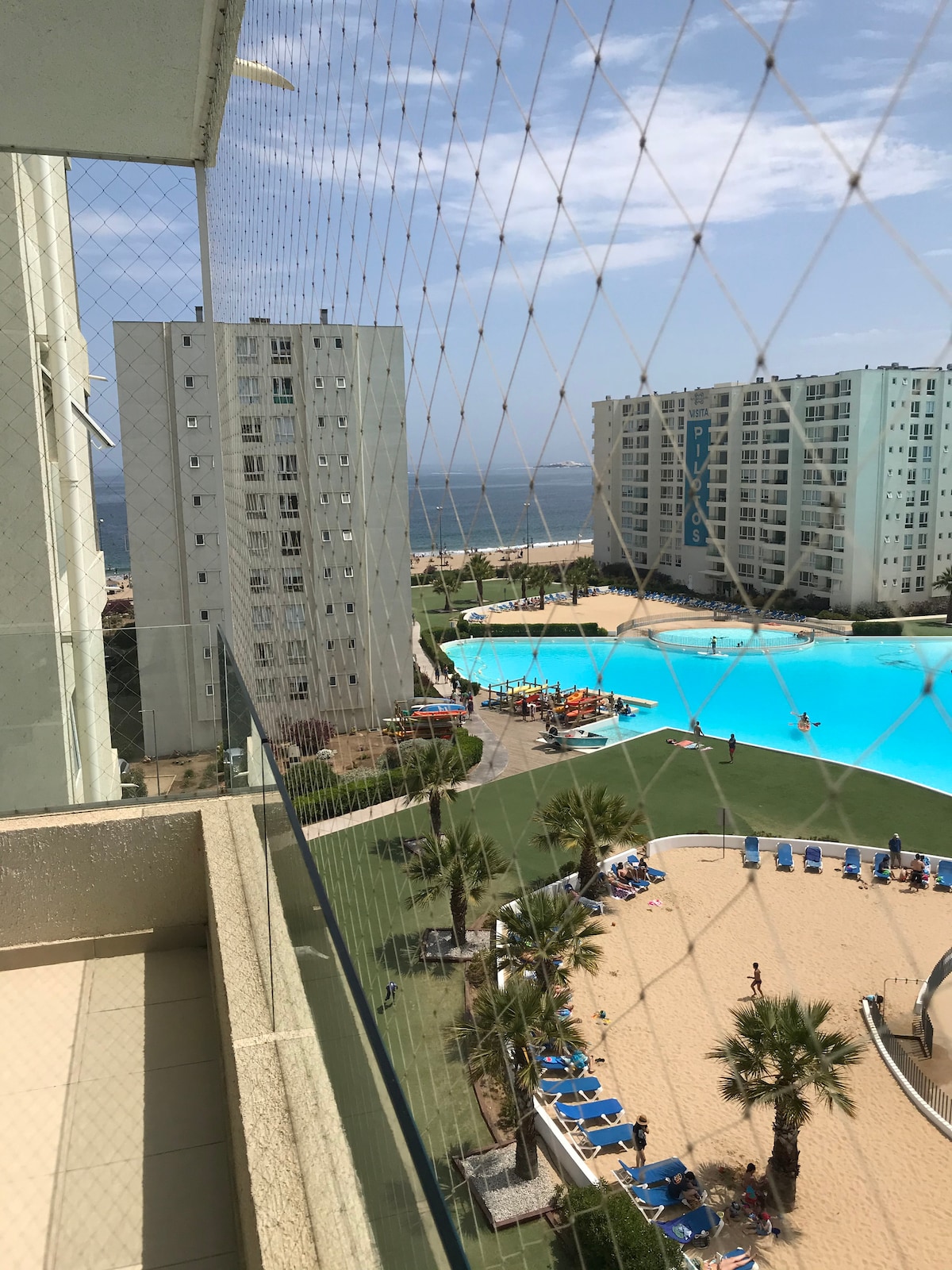 A view from the eighth floor shows a large pool area surrounded by sandy beach and palm trees. The ocean can be seen in the distance, with sun loungers arranged neatly around the pool. A safety netting is visible in the foreground.