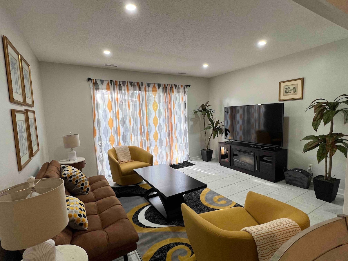 The living area features a comfortable brown sofa and two yellow chairs, arranged around a dark coffee table. A large television is positioned on a low stand, while the space is enhanced by decorative plants and bright patterned curtains that frame the window.
